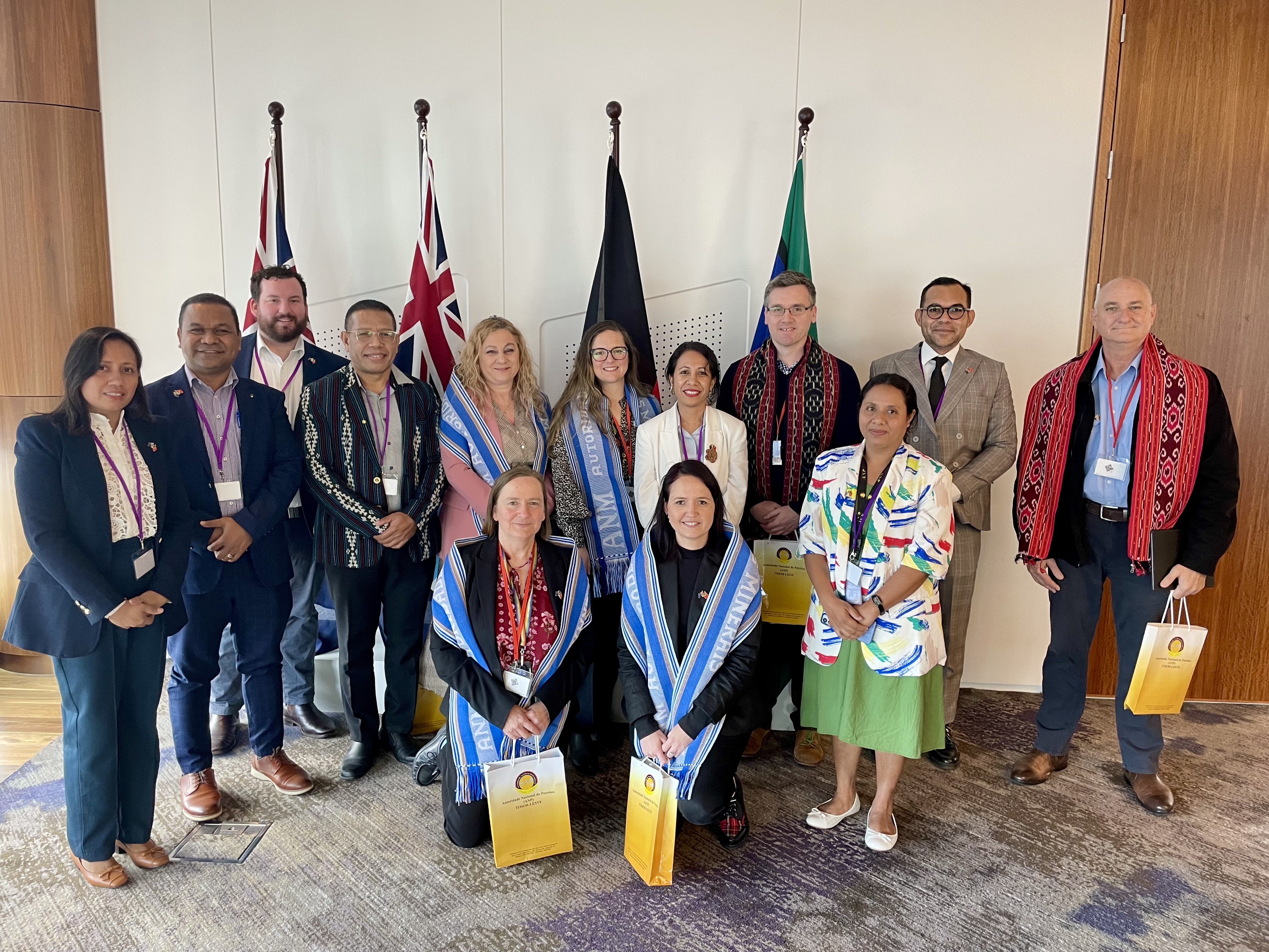 Photograph shows a group of delegates posing indoors for a formal gathering, with ceremonial flags behind them. Participants wear conference lanyards, patterned clothing, traditional garments, and colorful accessories, while several hold yellow gift bags.