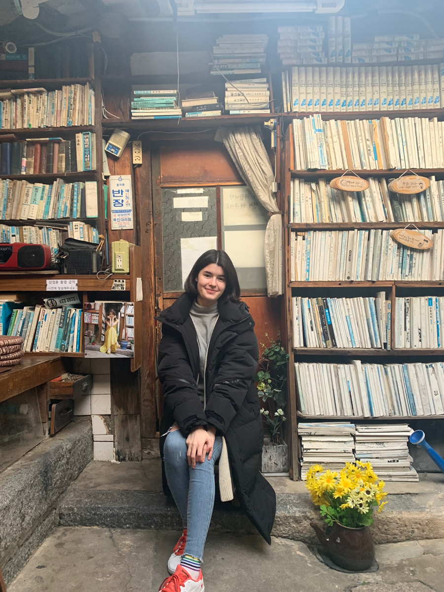 Photo of Seana sitting in front several bookshelves. 