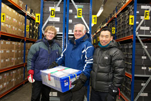Three people, one holding a box, standing at the end of long and high rows of storage shelves, full of boxes.