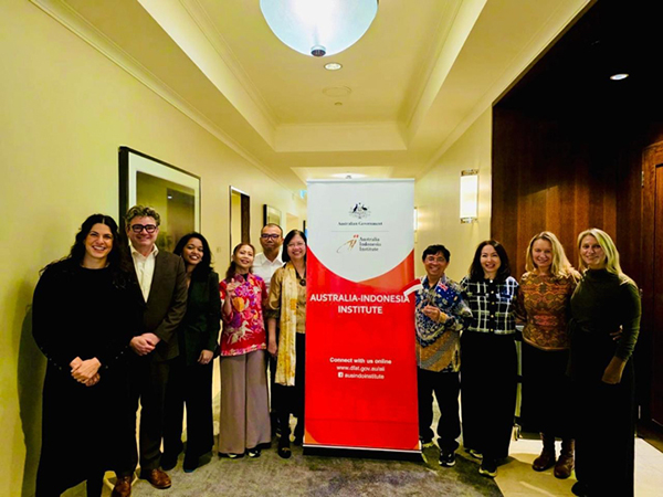 Group of about ten people standing in a hallway beside a red “Australia‑Indonesia Institute” banner, posing in a line against light-colored walls.