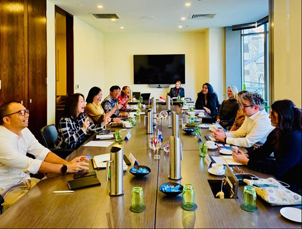 Long conference table meeting with about a dozen participants seated on both sides, notebooks and drinks on table, large screen at end, bright room with windows and wood panels.