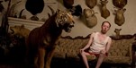 Man on the couch, in singlet and boxers, shying away from a stuffed tiger with antelope heads mounted on shields on wall behind him
