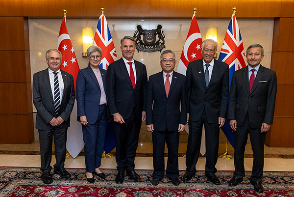 Six individuals in formal attire standing in front of Singapore and United Kingdom flags, suggesting a diplomatic or official event, with an emblem and lights in the background enhancing the formal setting