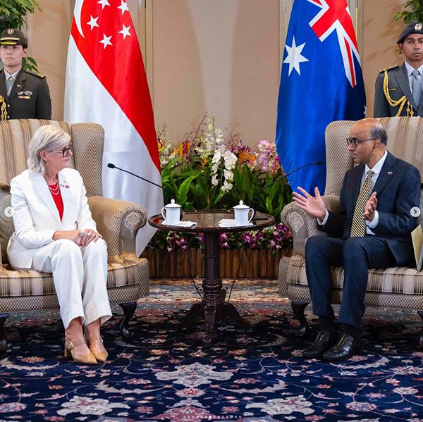 wo individuals seated in armchairs engaged in a formal conversation, with Singapore and Australia flags behind them, a table with teacups between them, and two uniformed personnel standing in the background.