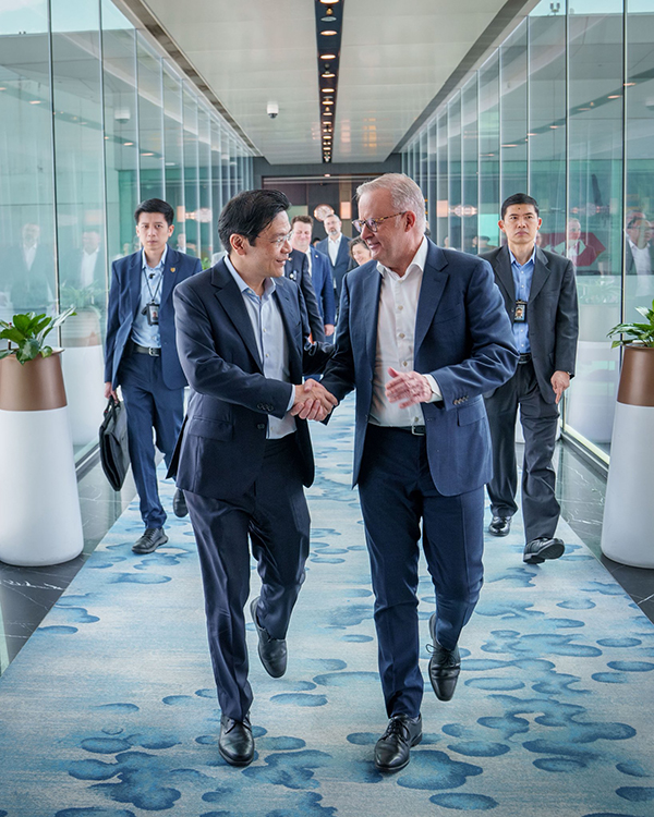 Two individuals in business attire shaking hands while walking down a well-lit hallway with glass walls, accompanied by others in similar attire, suggesting a formal meeting or diplomatic agreement