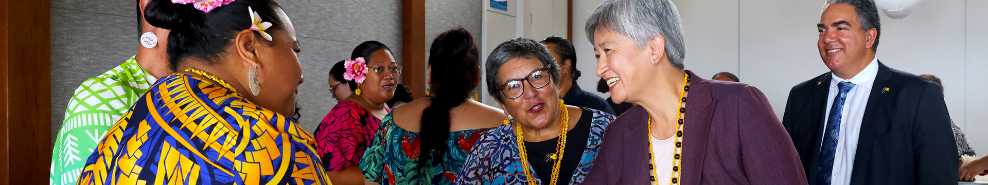 A group of people are gathered indoors, standing close together and interacting. They are wearing bright, patterned clothing and floral or beaded accessories. Several individuals wear colorful dresses with bold designs, while another wears a dark jacket with a long yellow beaded necklace. The setting appears to be a social or cultural event, with people positioned near a wall and doorway in a well-lit room.