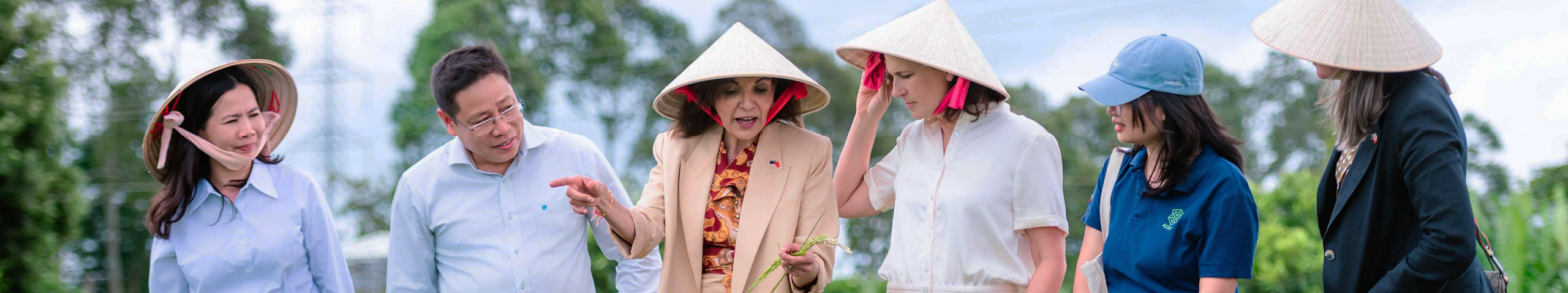 Minister for International Development The Hon Dr Anne Aly MP and people wearing traditional conical hats stand in a rice field, discussing and examining crops with local representatives.