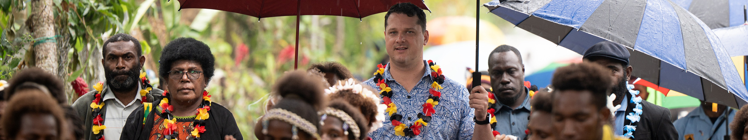 Community members and visitors stand under umbrellas in the rain while dancers in traditional dress perform in the foreground.