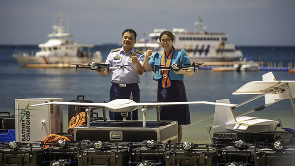 Two individuals standing on a dock display marine drones and equipment. They each hold small drone devices while larger aerial and underwater drones sit on cases nearby. Boats and a ferry are visible on the sea in the background.
