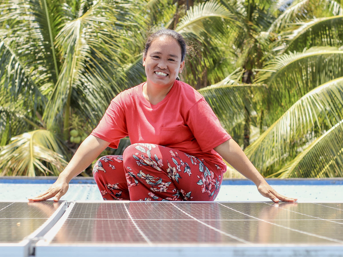 Photograph showing a smiling woman in a red shirt and patterned pants sitting behind a large solar panel amid tropical palm trees. Blue-black photovoltaic grid and reflective surface highlight renewable-energy technology in a bright outdoor setting.