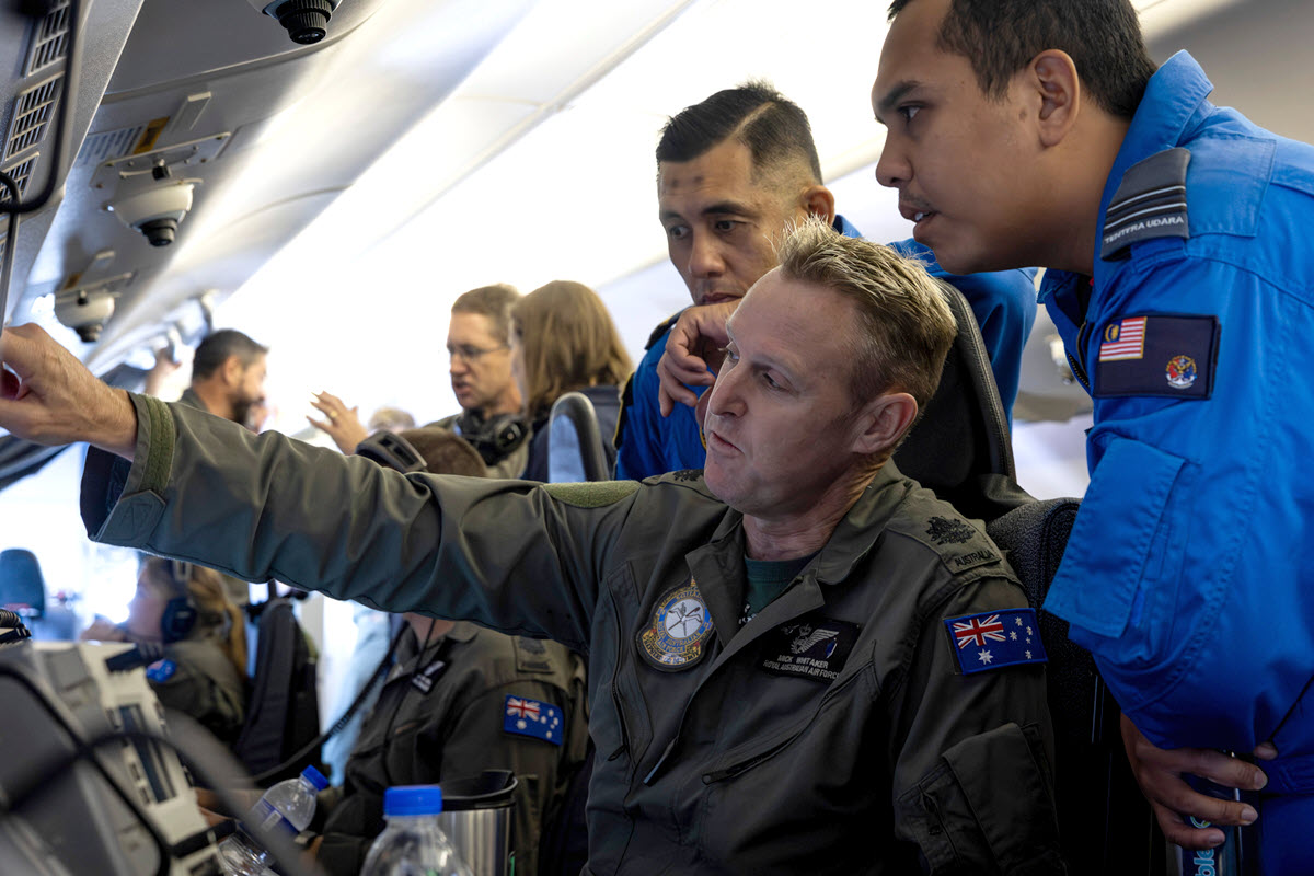 Photograph shows uniformed crew members inside an aircraft, with a seated man pointing toward equipment while colleagues observe. Blue and dark flight suits feature mission patches, highlighting an operational or training briefing in a crowded cabin.