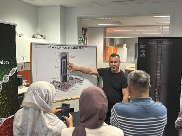 Presenter explains heavy ion accelerator technology to visitors using a facility diagram during an educational briefing and site visit.