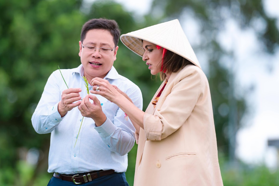 Photograph shows a man and woman examining a small green plant outdoors. Woman wears a traditional conical hat and beige coat, while man holds plant stem.