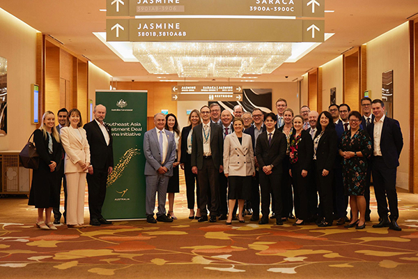 Group photo of approximately 25–30 attendees in business attire standing in a hotel conference hallway with warm lighting and patterned carpet. A green banner behind the group reads “Southeast Asia Investment Deal Roadshow" with an Australian emblem. The group faces the camera in a formal, staged arrangement.