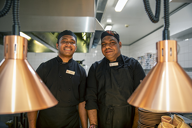 Two men dressed in chef’s uniform, in a kitchen, with heat lamps in the foreground.