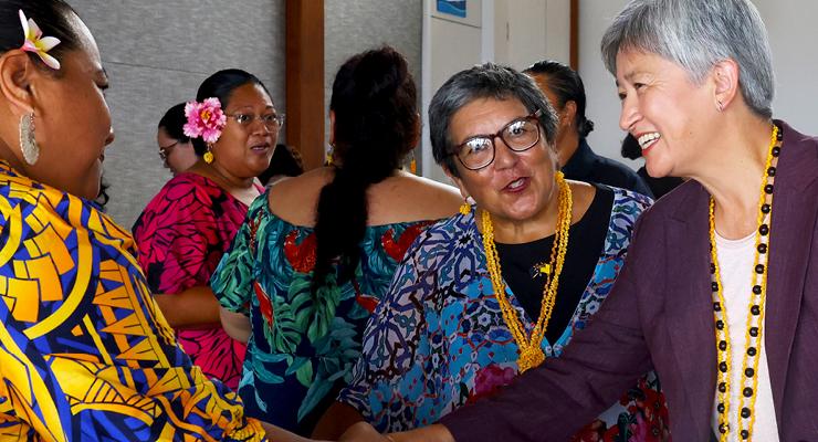 A group of people are gathered indoors, standing close together and interacting. They are wearing bright, patterned clothing and floral or beaded accessories. Several individuals wear colorful dresses with bold designs, while another wears a dark jacket with a long yellow beaded necklace. The setting appears to be a social or cultural event, with people positioned near a wall and doorway in a well-lit room.