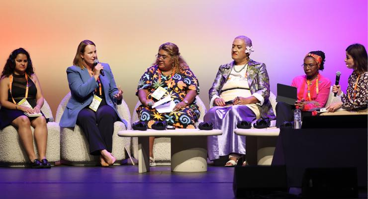 Panel speakers participate in a conference discussion on stage, seated in front of an audience during a formal event.