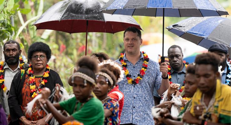 Community members and visitors stand under umbrellas in the rain while dancers in traditional dress perform in the foreground.