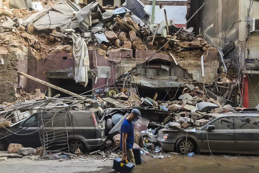 A man wearing a mask walks through the streets of Beirut, with damaged buildings and cars in the background.