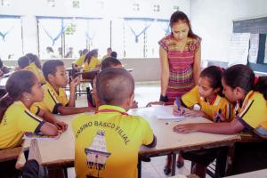 Six primary school students wearing school uniforms sit together at a table in a classroom working on a group learning activity. Their teacher stands next to the table smiling and looking at their work.