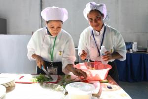 Two women hospitality students (in chef'attire) preparing food as part of the Timor-Leste's National Skills Competition. One woman is cutting vegetables for a salad and the other is preparing a sauce in a bowl.