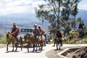 Four Timorese men riding Timorese ponies up a mountain road in Fatubosa, Aileu. They are using a road that has been recently rehabilitated. There is a four wheel drive vehicle behind them.