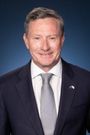 Photograph of a middle-aged man wearing a dark suit, white shirt, and light gray tie against a blue gradient background. The man has short hair and a small lapel pin on his left side, smiling at the camera.