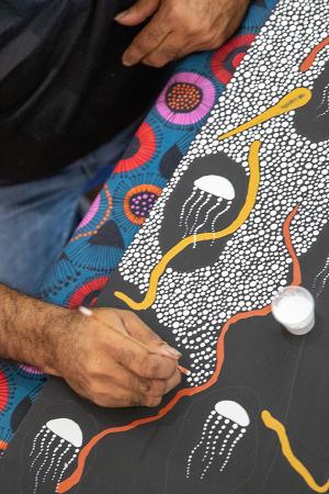 Close-up of a hand painting an Aboriginal dot artwork featuring white jellyfish shapes, orange and yellow lines, and intricate patterns.