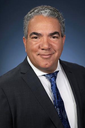 Paul Harrington, smiling, wearing a black blazer, white collared shirt and a blue tie.