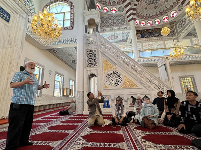 Group of people seated on a carpet listening to a religious leader speaking in a mosque.