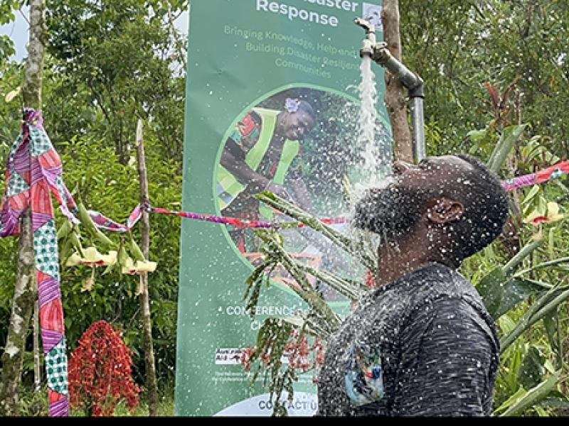 Person stands outdoors under a flowing pipe, rinsing with water amid lush greenery, while a colorful decorated area and a “Disaster Response” sign highlight a community resilience or hygiene awareness event.
