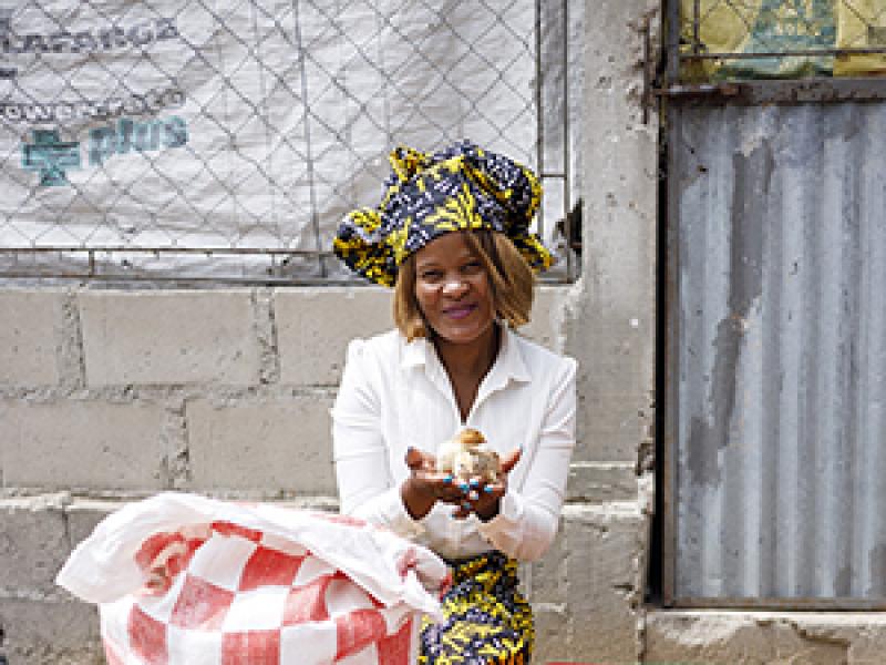 A person kneels on the ground holding a small chicken, surrounded by poultry farming supplies including a checkered bag, plastic feeders, and a ventilated crate, with a concrete wall and corrugated metal sheet in the background.