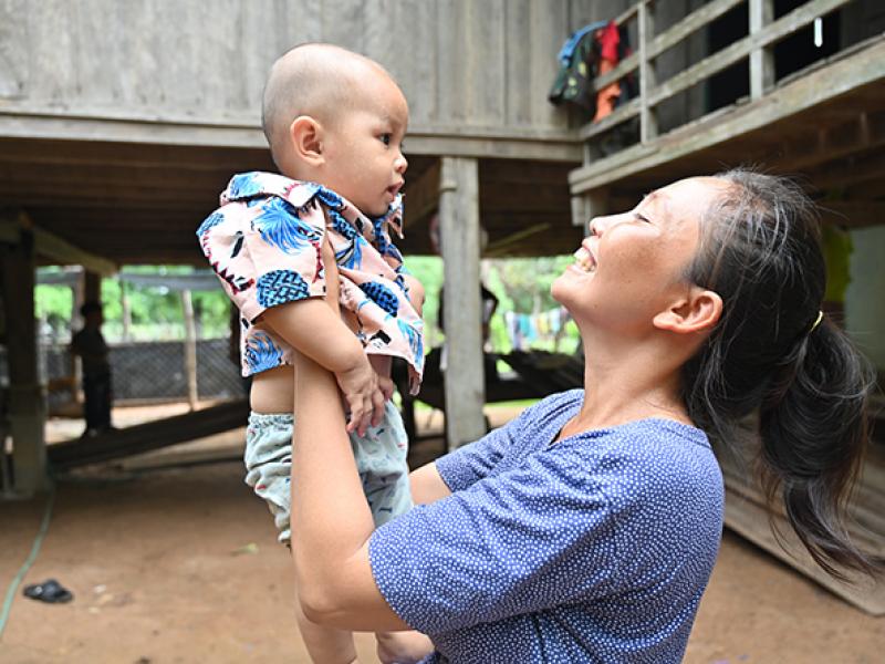 A person is standing outdoors under a raised wooden structure, holding up a small child at arm’s length. The child is wearing a patterned shirt and light-colored shorts. The setting includes wooden posts, a dirt ground, and parts of a building with an elevated walkway and railing in the background.