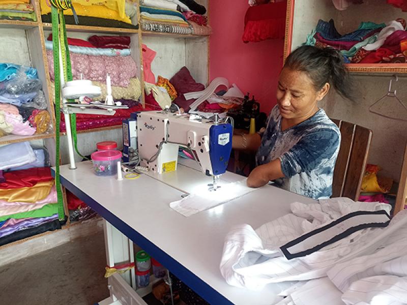 A person sits at a sewing workstation, operating an industrial sewing machine. Shelves behind the workspace are stacked with colorful folded fabrics, and various sewing supplies and materials are arranged on the table. A partially completed garment lies in front of the machine.