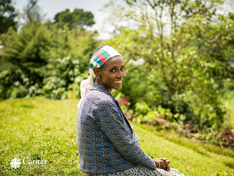 A person is sitting on a grassy hillside in an outdoor setting, wearing a patterned blue jacket and a headscarf with green, white, and red stripes. The background features lush greenery, trees, and a slightly blurred landscape, suggesting a rural or natural environment. The Caritas Australia logo is visible in the bottom left corner of the image.