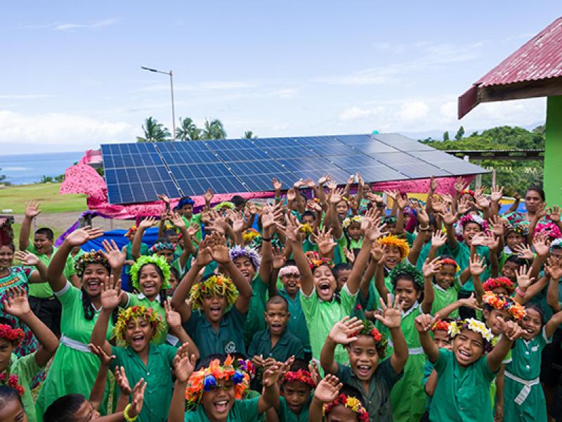 A large group of children wearing matching green clothing and colorful flower garlands raise their hands while standing outdoors. Behind the group is a structure with a rooftop covered in solar panels. The scene is bright and tropical, with palm trees, green grass, and the ocean visible in the background.