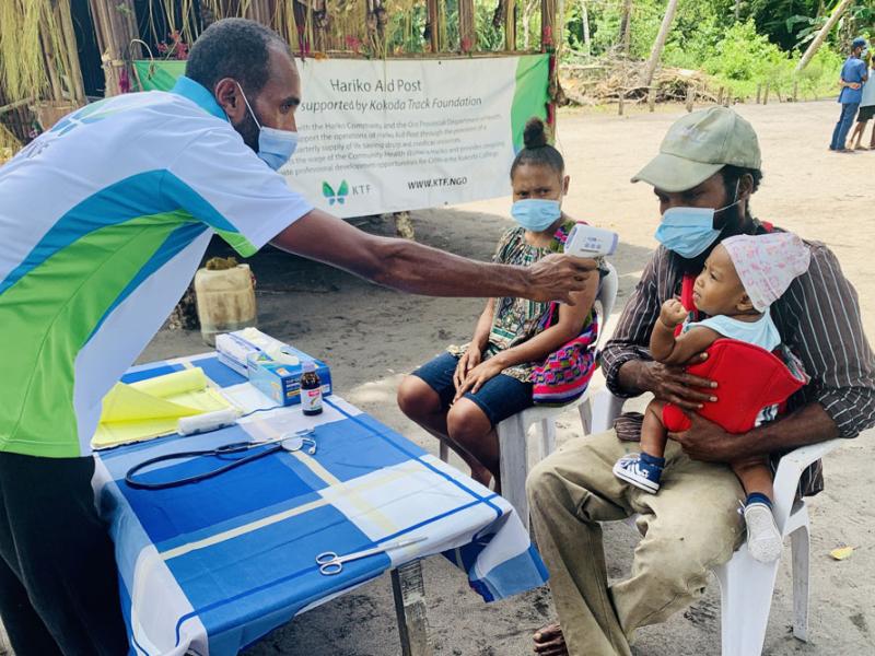 Community Health Worker doing a temperature check on a baby sitting in the fathers lap. 