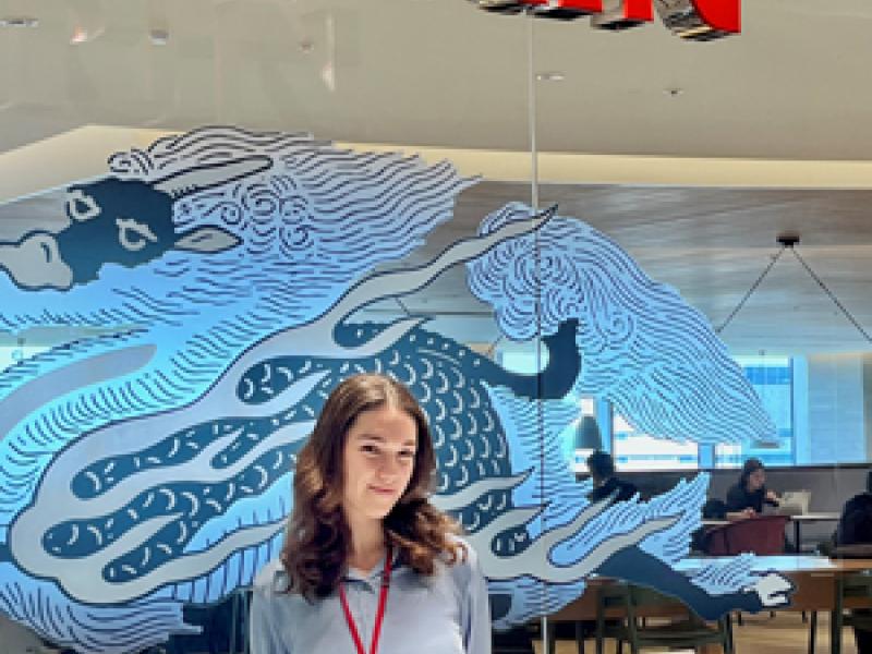 A participant stands at the reception area of a Kirin office. A large red Kirin sign hangs above, with decorative artwork on a glass wall and a reception desk in the foreground.