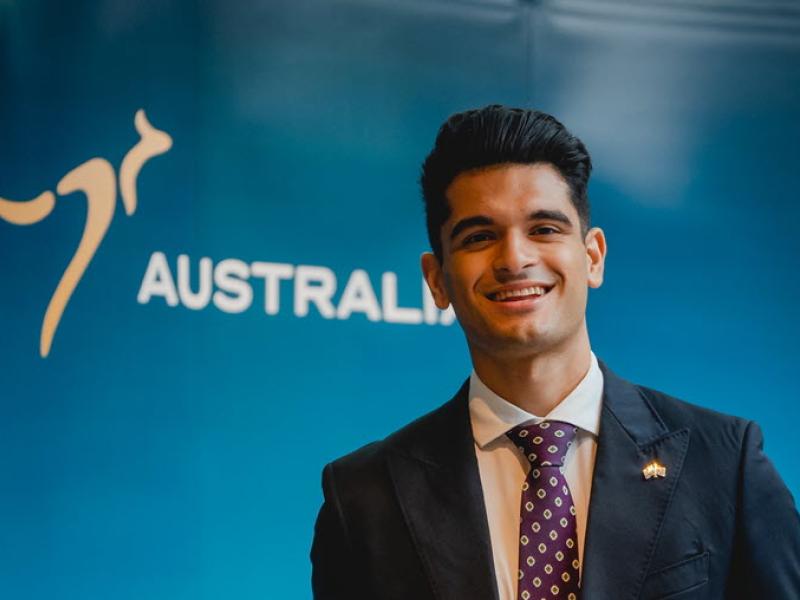 Ricky standing in front of an 'Australia' nation brand banner, dressed in a dark blue suit, white business shirt, and a maroon tie with white chevron pattern. 