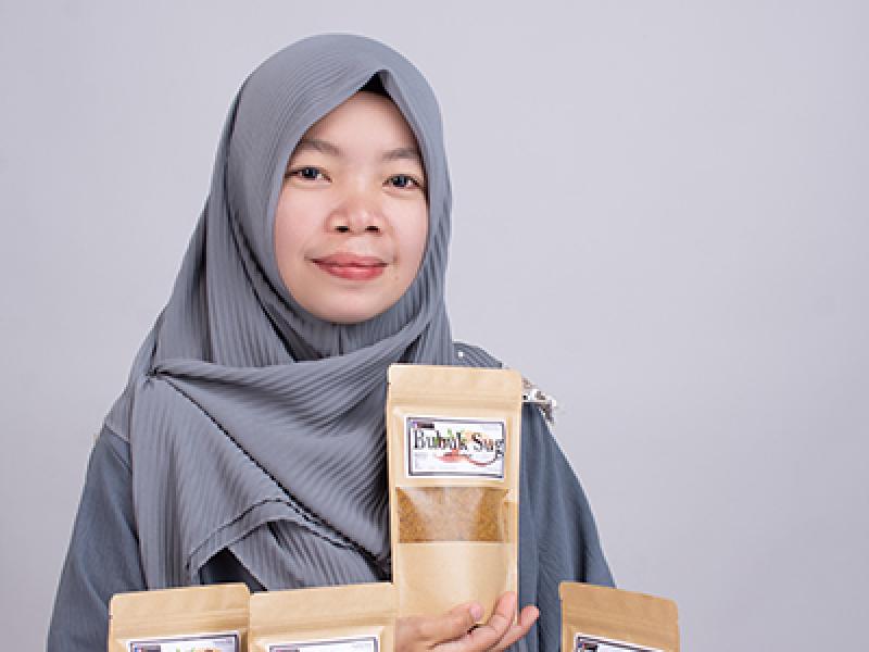 A woman wearing a hijab holds a packed of bobuk, with more packages on display
