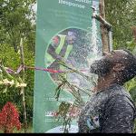 Person stands outdoors under a flowing pipe, rinsing with water amid lush greenery, while a colorful decorated area and a “Disaster Response” sign highlight a community resilience or hygiene awareness event.