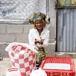 A person kneels on the ground holding a small chicken, surrounded by poultry farming supplies including a checkered bag, plastic feeders, and a ventilated crate, with a concrete wall and corrugated metal sheet in the background.