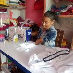 A person sits at a sewing workstation, operating an industrial sewing machine. Shelves behind the workspace are stacked with colorful folded fabrics, and various sewing supplies and materials are arranged on the table. A partially completed garment lies in front of the machine.