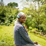 A person is sitting on a grassy hillside in an outdoor setting, wearing a patterned blue jacket and a headscarf with green, white, and red stripes. The background features lush greenery, trees, and a slightly blurred landscape, suggesting a rural or natural environment. The Caritas Australia logo is visible in the bottom left corner of the image.