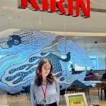 A participant stands at the reception area of a Kirin office. A large red Kirin sign hangs above, with decorative artwork on a glass wall and a reception desk in the foreground.
