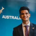 Ricky standing in front of an 'Australia' nation brand banner, dressed in a dark blue suit, white business shirt, and a maroon tie with white chevron pattern. 
