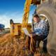 Photo of a man sitting in a large tractor tire with a laptop on his lap in a large farm field. Tractor plowing in the background.