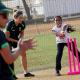 A young Indian wicketkeeper, arms outstretched, braces to catch a cricket ball. She is cheered on by two young Australian women cricketers.