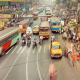 A busy shopping street, crowded with pedestrians. Road traffic consists predominantly of buses, motorcycles and some taxis.