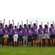Group of young women facing away from the photographer all pumping a fist into the air and wearing their purple matching shirts. 